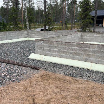 Betoniset tukimuurit ja sorapolku metsäisellä ulkoalueella, talo osittain näkyvissä oikealla.