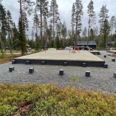 Ulkona soraleikkikenttä, jossa korotettu hiekkalaatikko, hajallaan olevia palikoita ja taustalla korkeita mäntyjä