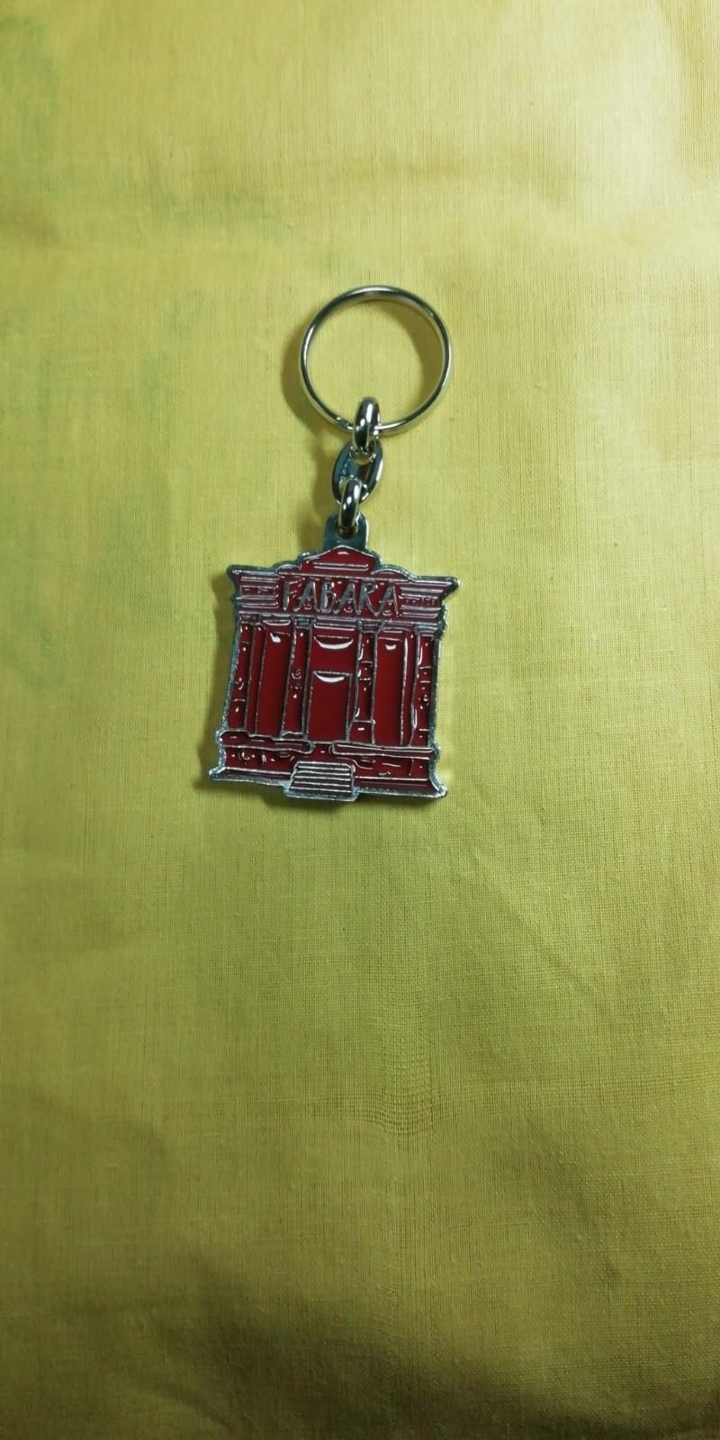 Llavero de metal de color rojo y plata con diseño de edificio, sobre fondo de tela de color amarillo.
