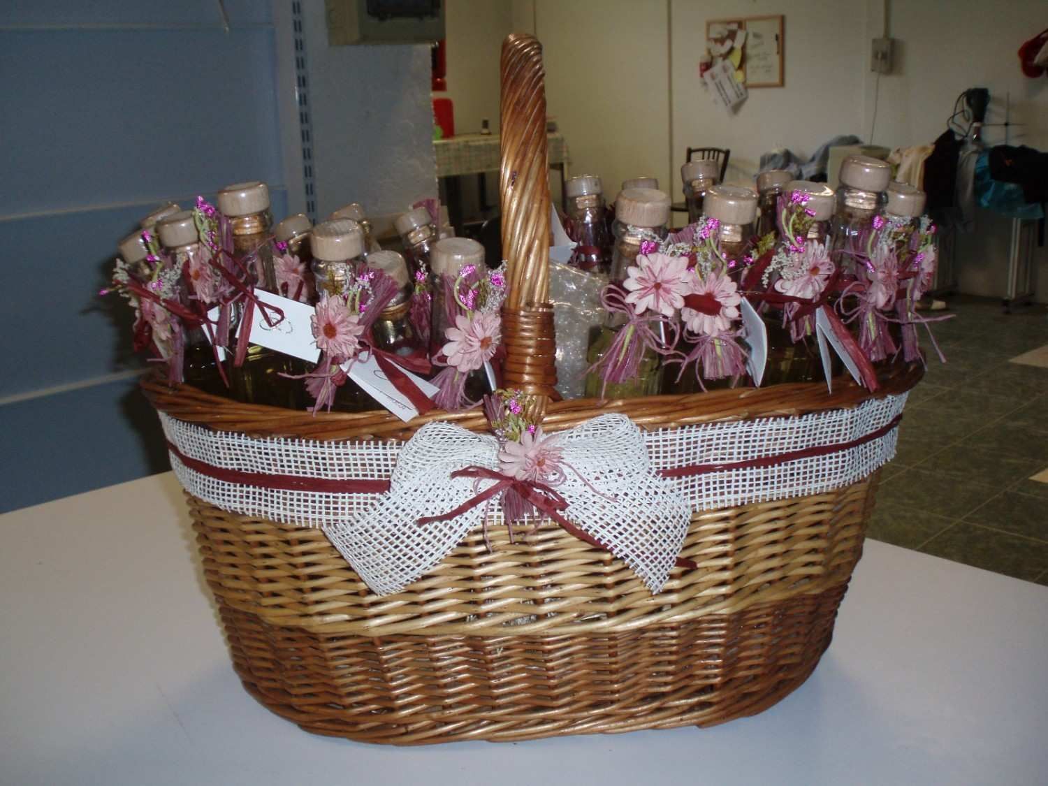 Una cesta de mimbre llena de botellas de cristal, decorada con cintas y flores, reposa sobre una superficie blanca.