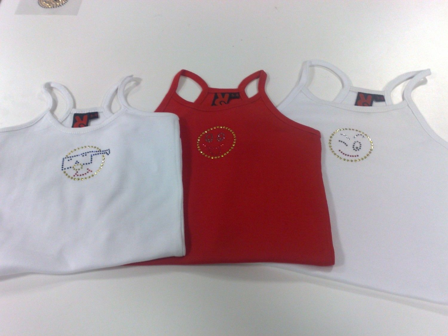 Tres camisetas sin mangas: dos blancas y una roja, con diseños bordados en dorado.