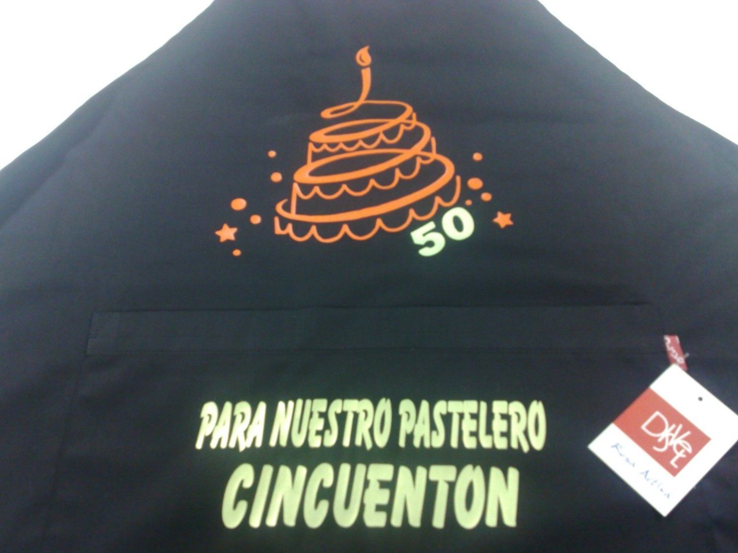 Delantal negro con diseño de pastel naranja y texto en español: 