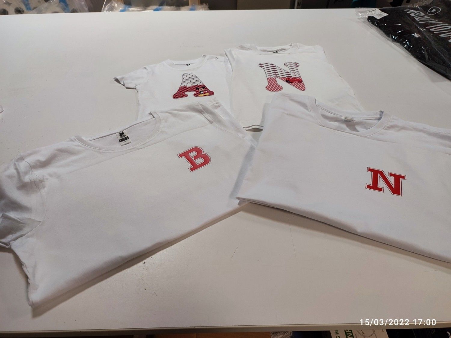 Camisetas blancas con letras rojas bordadas A, B y N dispuestas sobre una superficie blanca.
