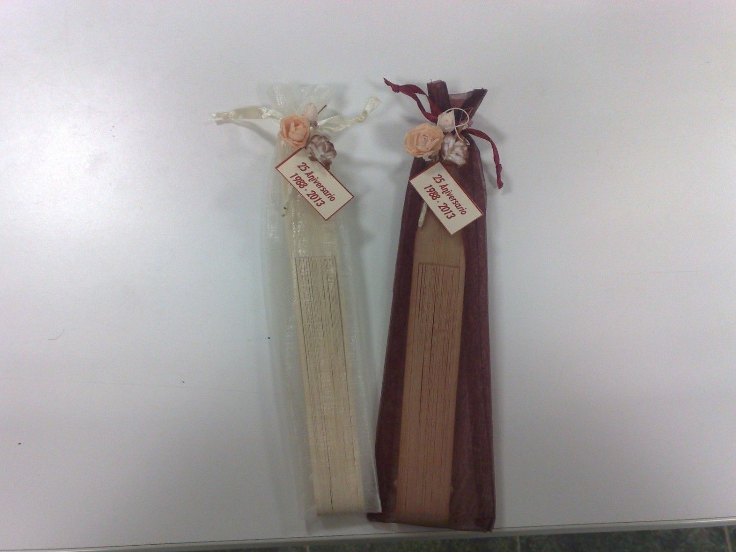 Dos abanicos decorativos, uno beige y otro marrón, en bolsas de tela con etiquetas y detalles florales.