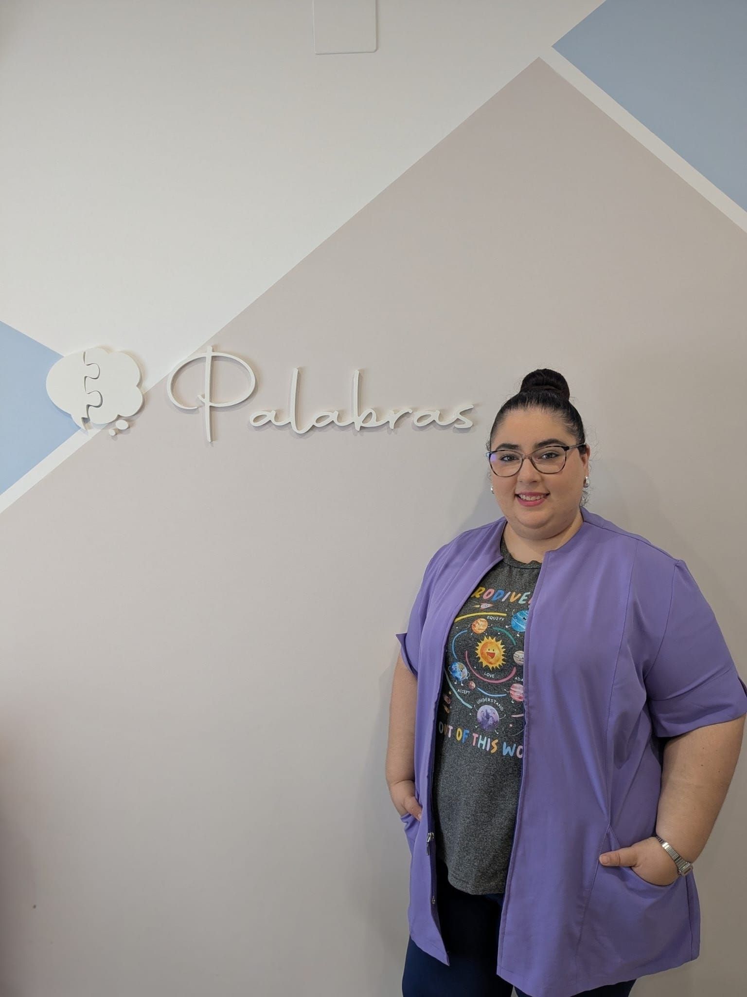 Una mujer vestida con un uniforme médico morado está de pie junto a una pared con la palabra 