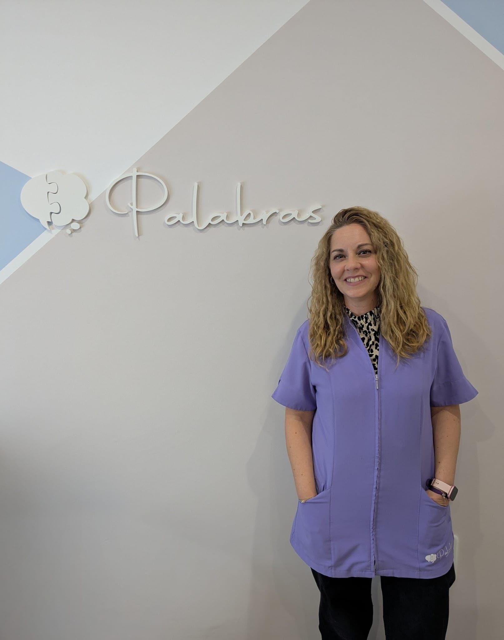 Una mujer vestida con un uniforme médico morado está de pie junto a una pared con la palabra 
