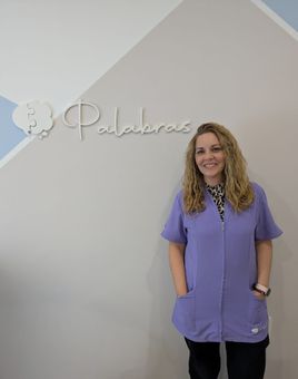 Una mujer vestida con un uniforme médico morado está de pie junto a una pared con la palabra