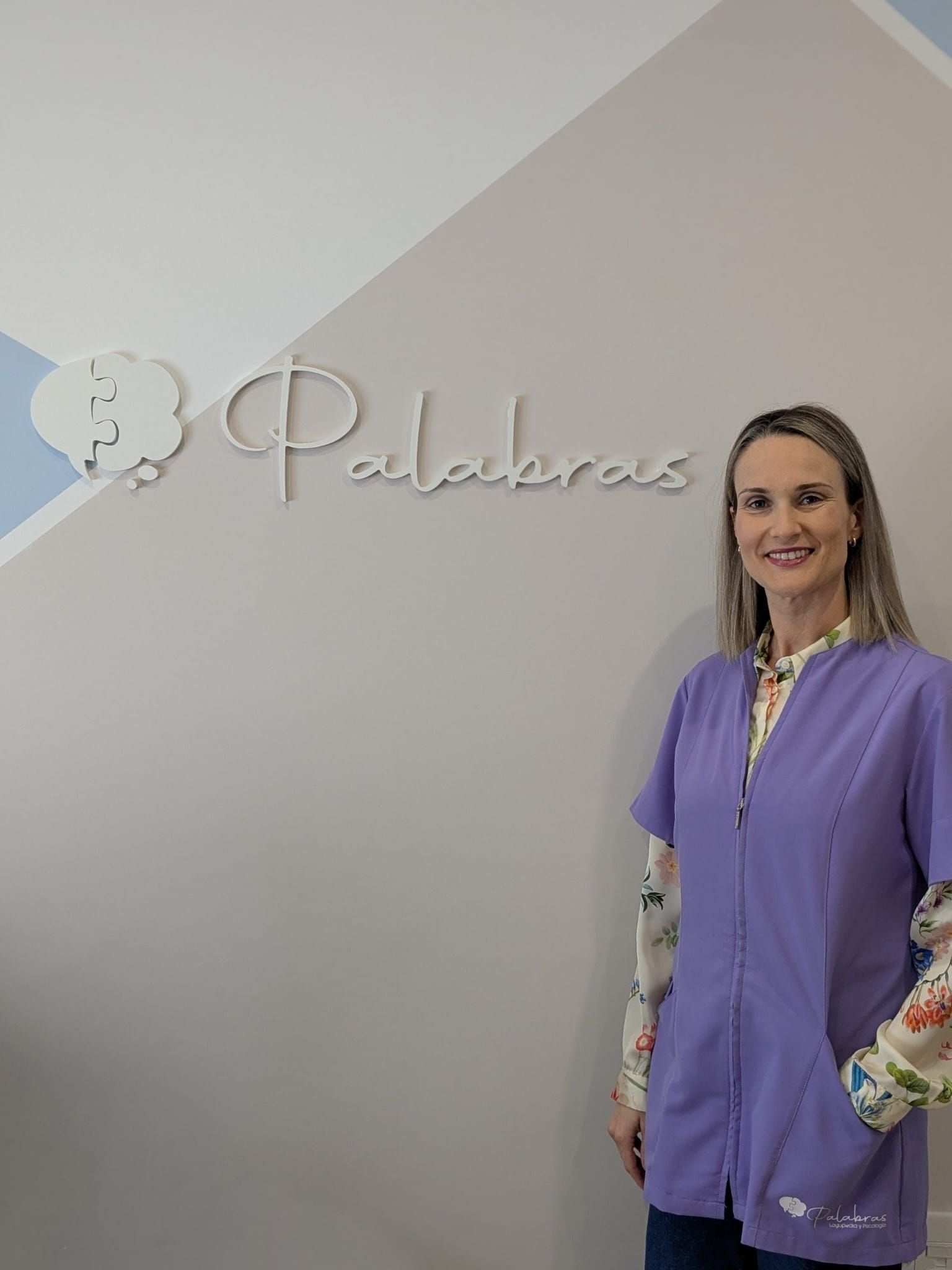 Una mujer con una chaqueta morada está de pie frente a una pared con un logotipo que dice 