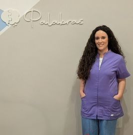 Una mujer con uniforme lavanda sonríe, de pie junto al logotipo de