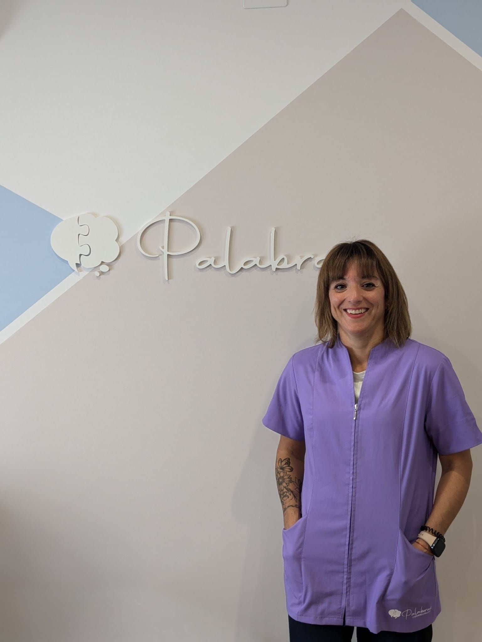 Una mujer con uniforme lavanda sonríe, de pie junto al logotipo de 