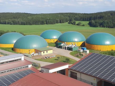 Fünf türkisfarbene Biogaskuppeln mit gelben Sockeln auf einem Feld, im Vordergrund ein Gebäude und Solarmodule.