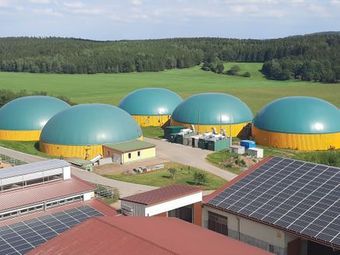 Fünf türkisfarbene Biogaskuppeln mit gelben Sockeln auf einem Feld, im Vordergrund ein Gebäude und Solarmodule.