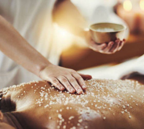 Une personne reçoit un soin spa avec un gommage appliqué sur le dos tandis qu'un praticien tient un bol d'ingrédients.