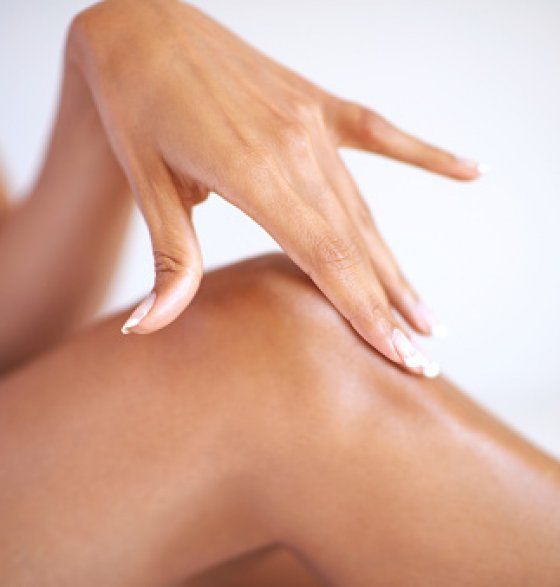 Une main effleure délicatement un genou lisse et nu et y applique une crème hydratante sur un fond clair et neutre.
