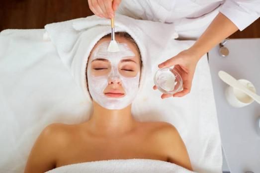 Une personne est allongée sur un lit de spa, les yeux fermés, et reçoit un masque facial appliqué par un praticien.