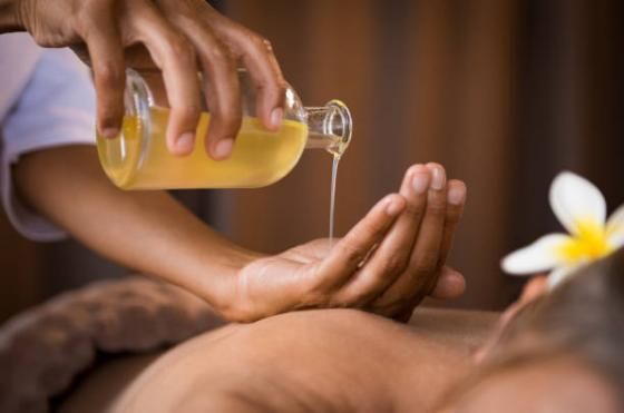 Une personne verse de l'huile de massage dorée d'une bouteille en verre sur les mains d'une autre personne, au-dessus de celle-ci qui reçoit un soin au spa.