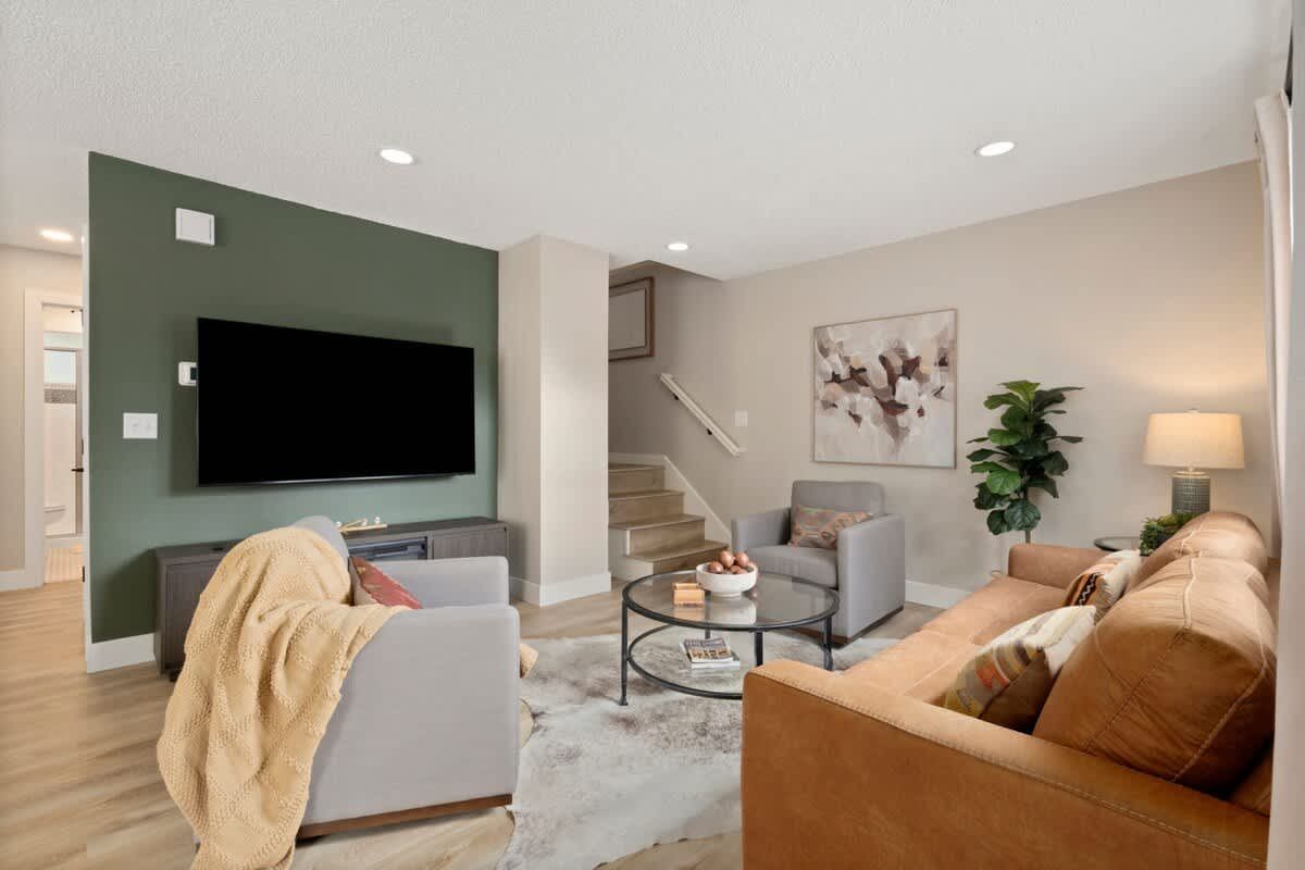 Modern living room with green accent wall, wall-mounted TV, tan sofa, armchair, and staircase.