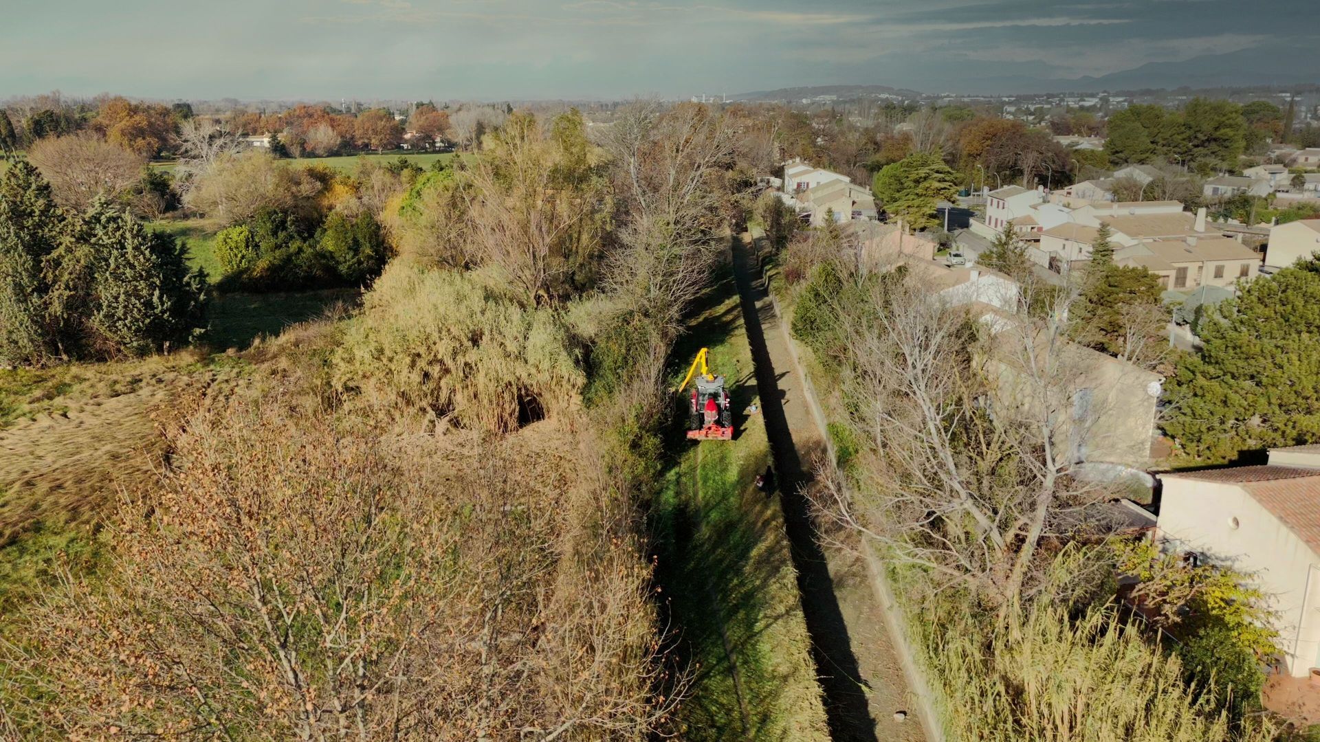 Vue aérienne d’un tracteur avec bras de débroussaillage en action pour nettoyer un canal bordé de végétation à Morières-lès-Avignon.