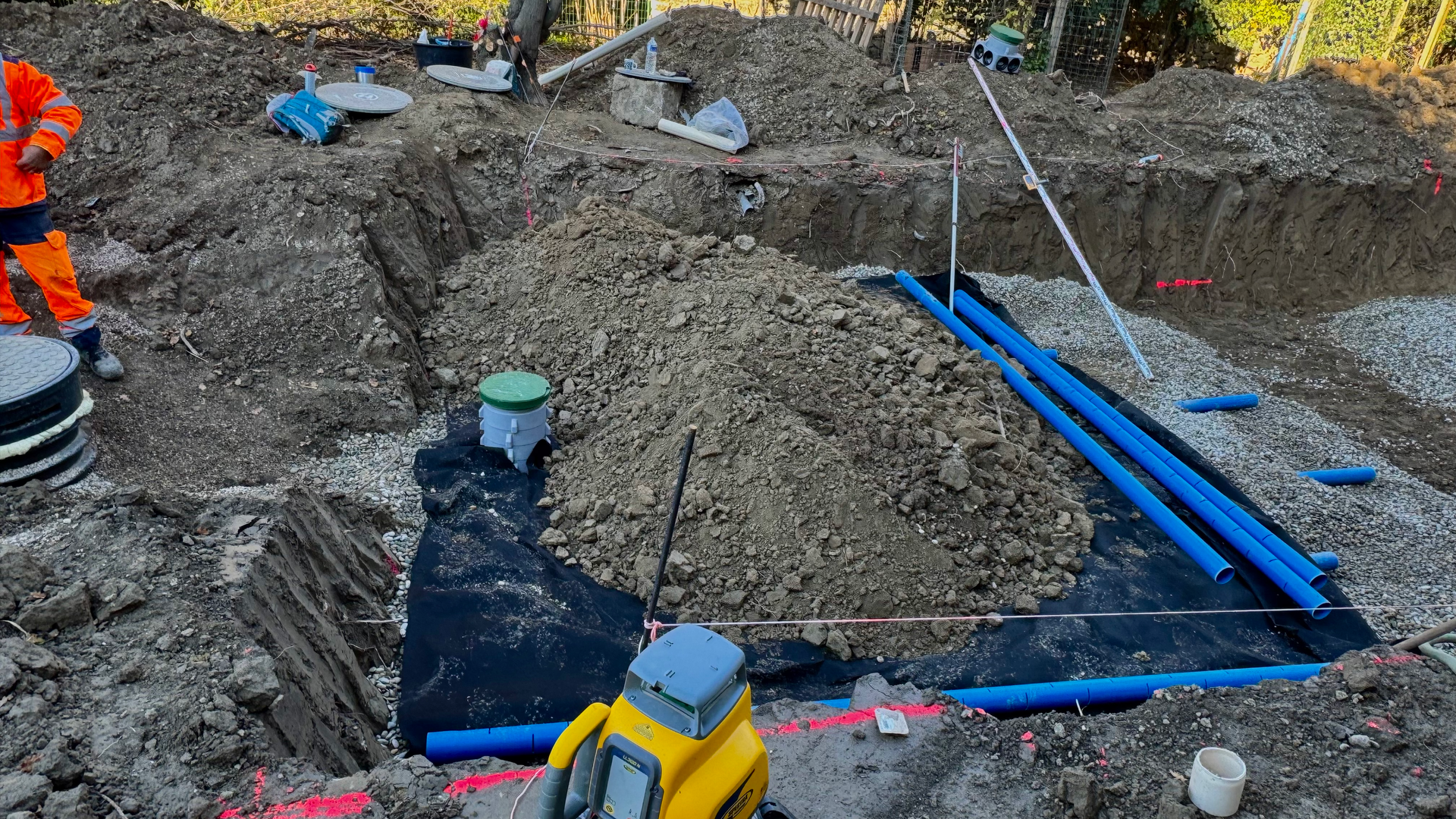 Vue détaillée d’un chantier d’assainissement avec des canalisations bleues en cours de pose, tranchées préparées et équipement de compactage du sol. Présence d’un regard d’inspection pour le réseau d’évacuation.