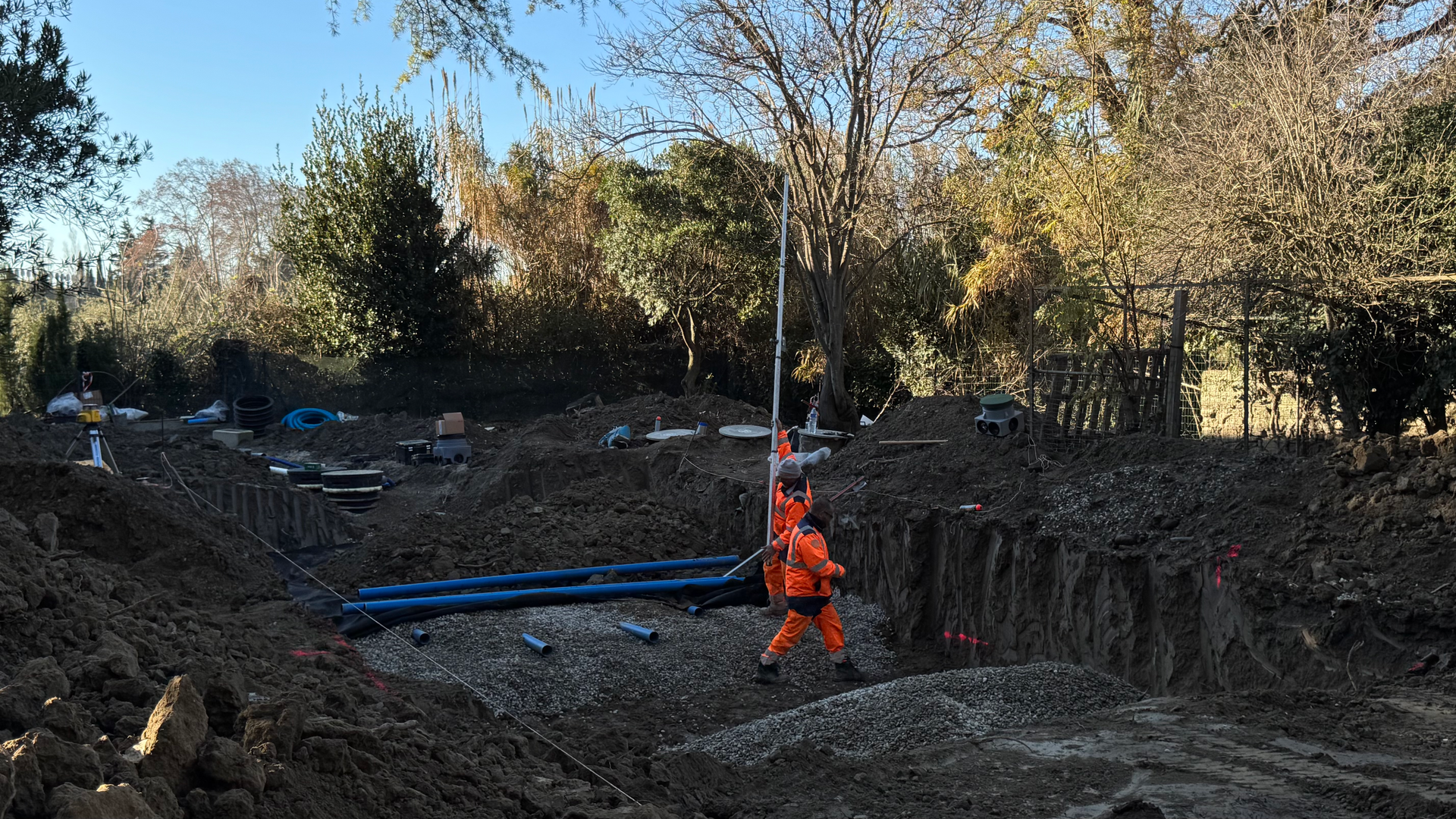 Chantier d’assainissement avec des ouvriers en tenue de sécurité posant des canalisations bleues dans une tranchée. Terrassement du terrain, préparation du lit de gravier et installation des équipements pour le réseau d’assainissement.