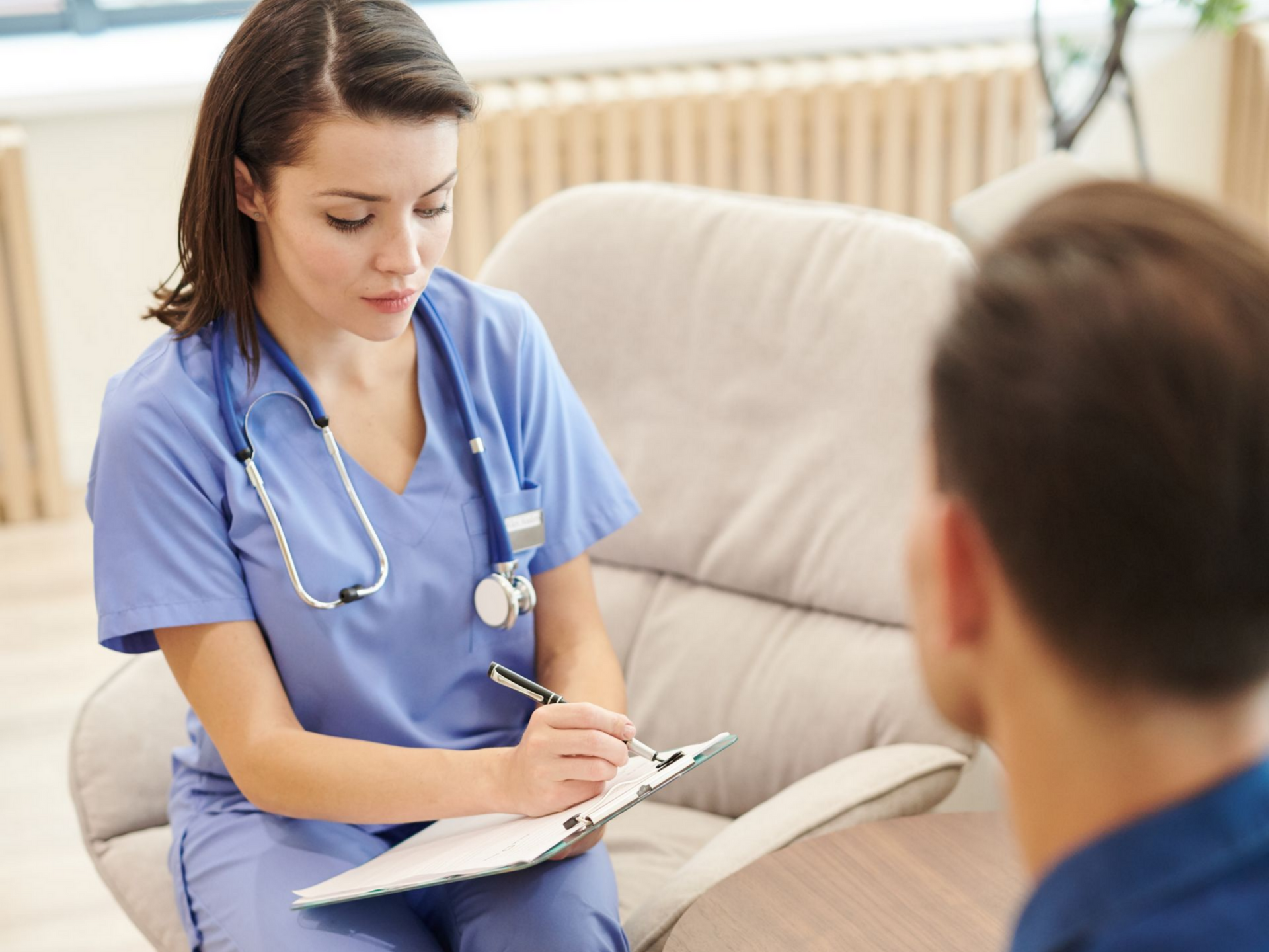 Une infirmière en blouse bleue prend des notes lors d'une consultation avec un patient.