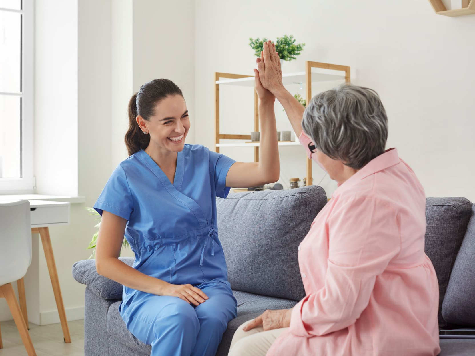 Une aide-soignante en blouse bleue tape dans la main d'une personne âgée assise sur un canapé.
