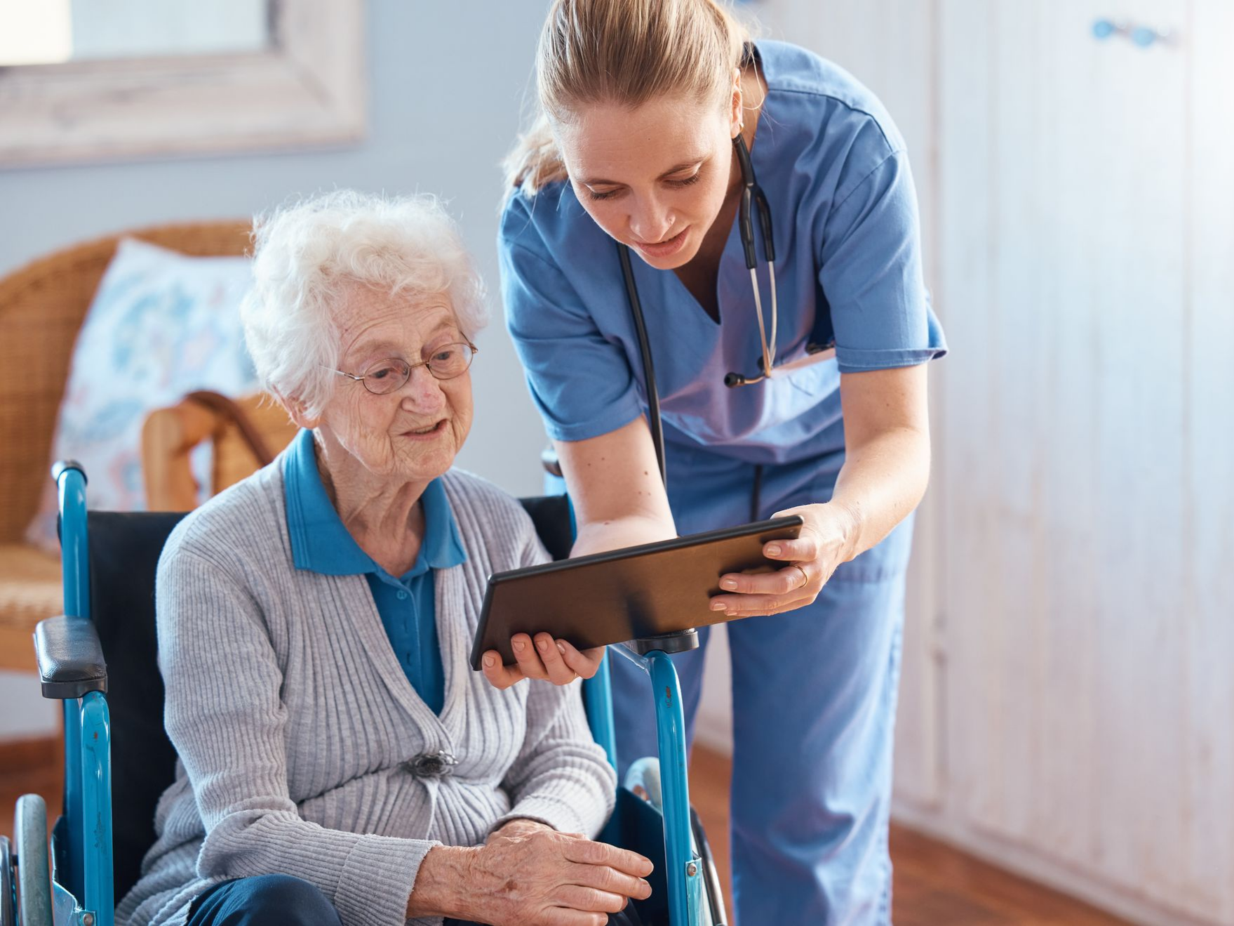 Une infirmière montre une tablette à une personne âgée en fauteuil roulant. Tous deux sont à l'intérieur et regardent l'écran.