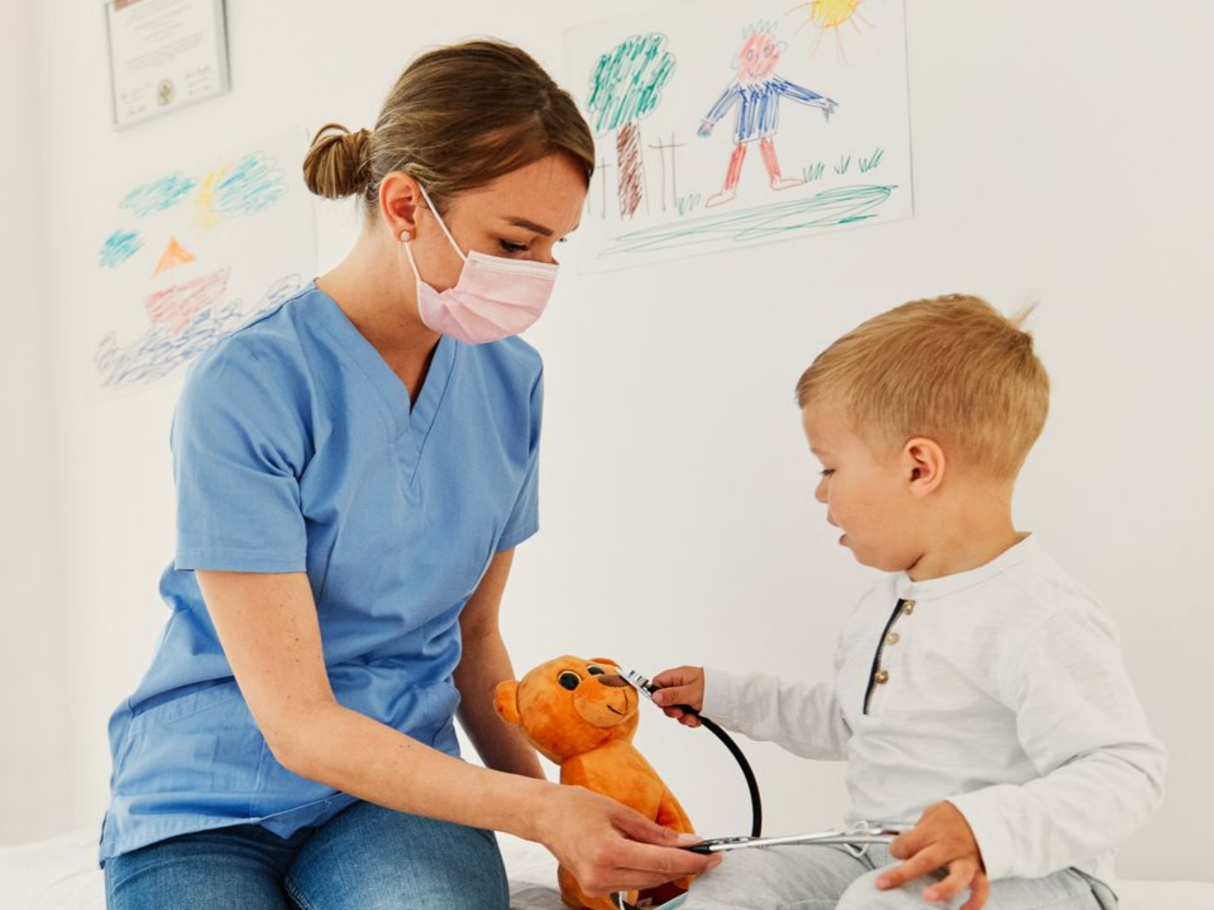 Une infirmière en blouse bleue utilise un stéthoscope sur un ours en peluche avec un enfant dans un contexte médical.