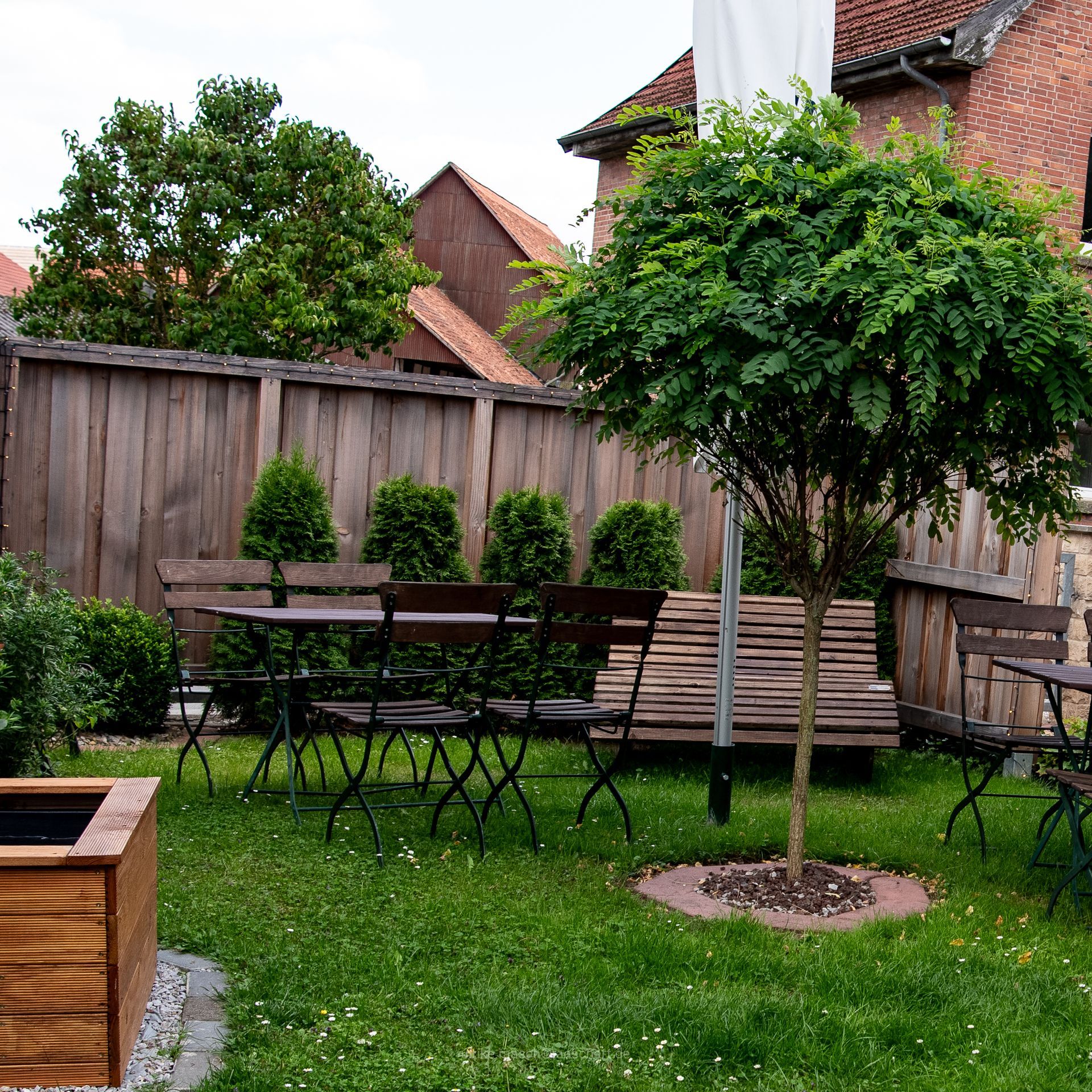 Sitzbereich im Freien mit Tischen, Stühlen, Baum und Holzzaun. Grünes Gras und kleine Pflanzen.