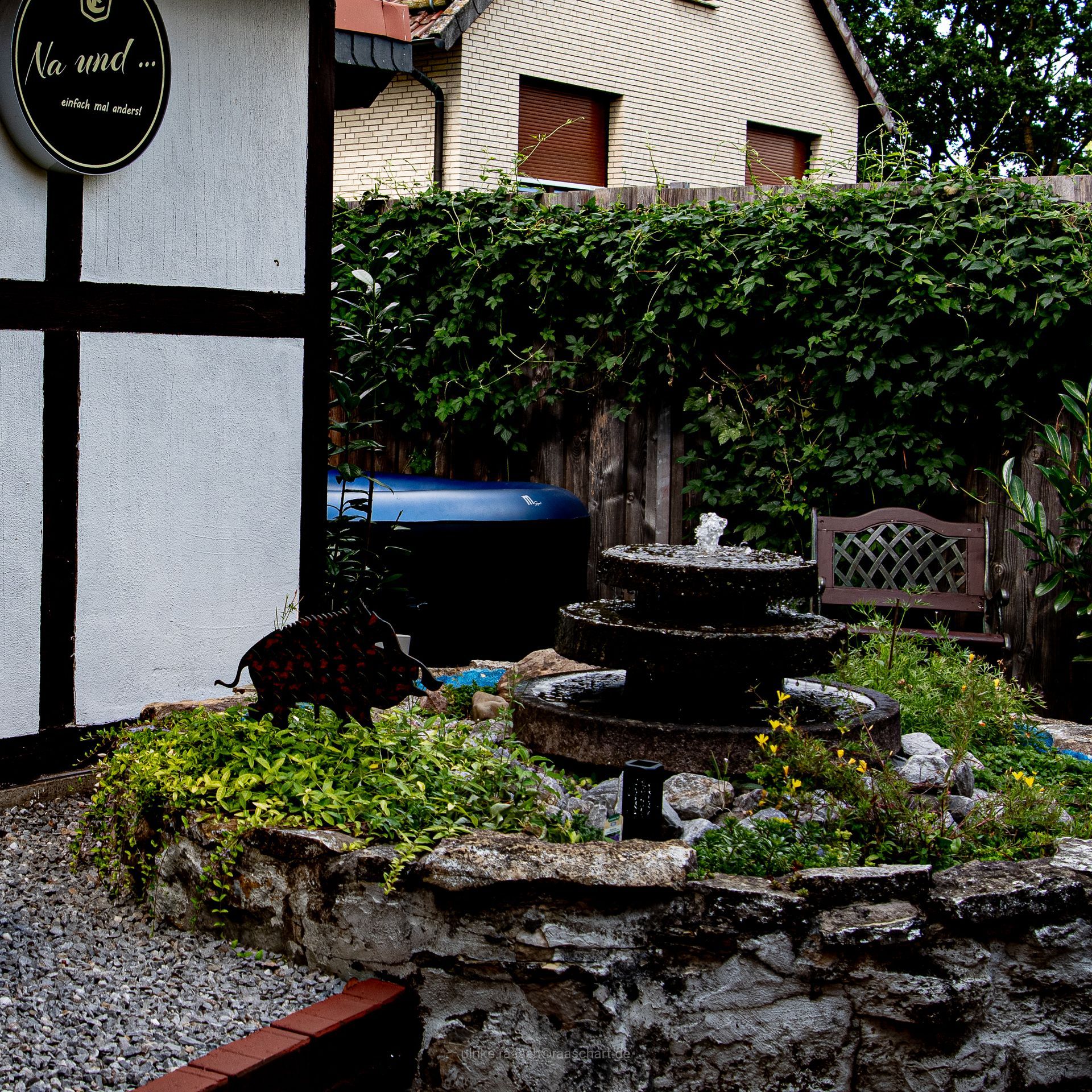 Steinbrunnen in einem Garten. Efeubewachsener Zaun und Haus im Hintergrund. Weiße Gebäudefassade mit schwarzen Balken links.
