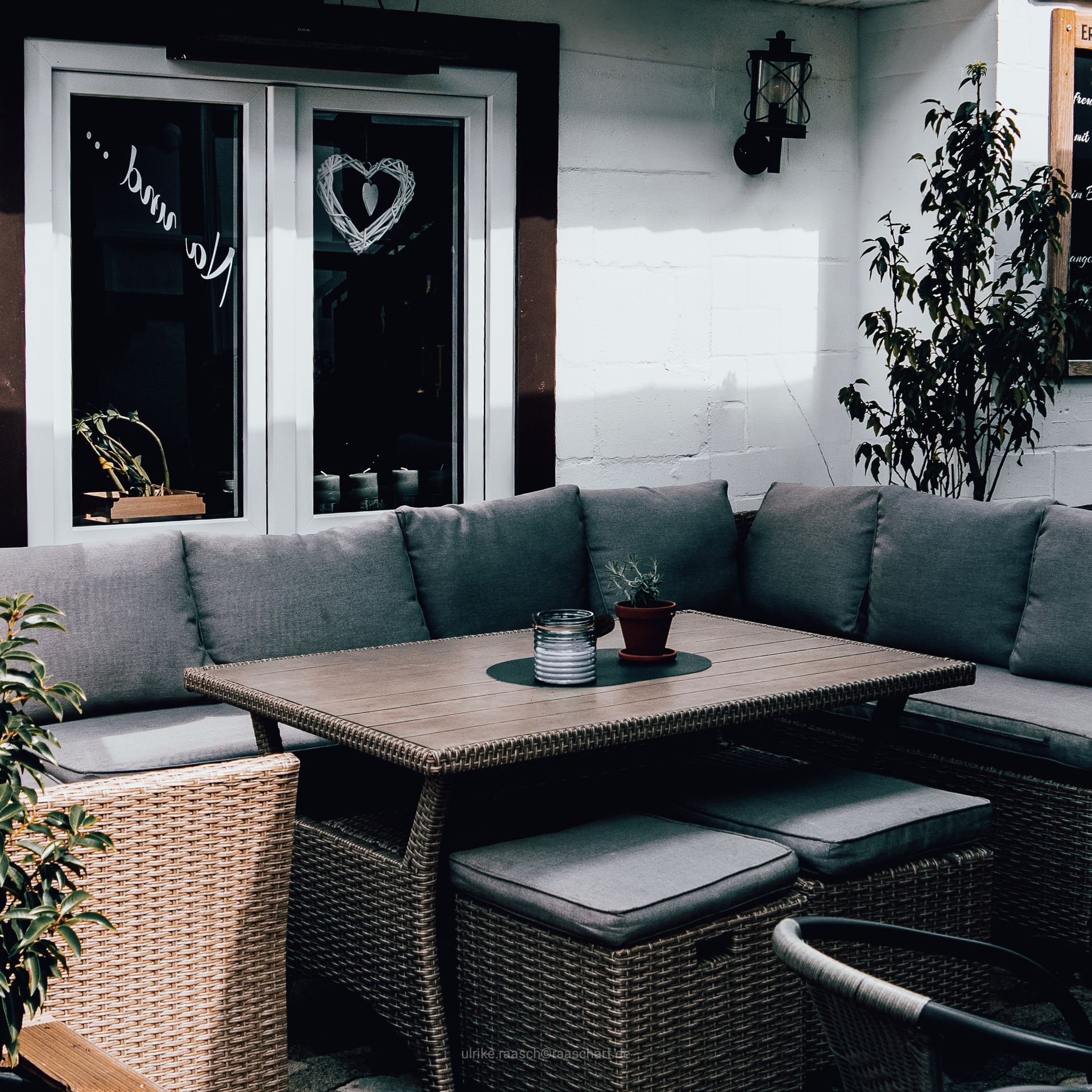 Sitzecke im Freien mit Korbmöbelsofa, Tisch und Kissen. Im Hintergrund ein Fenster mit herzförmiger Dekoration.