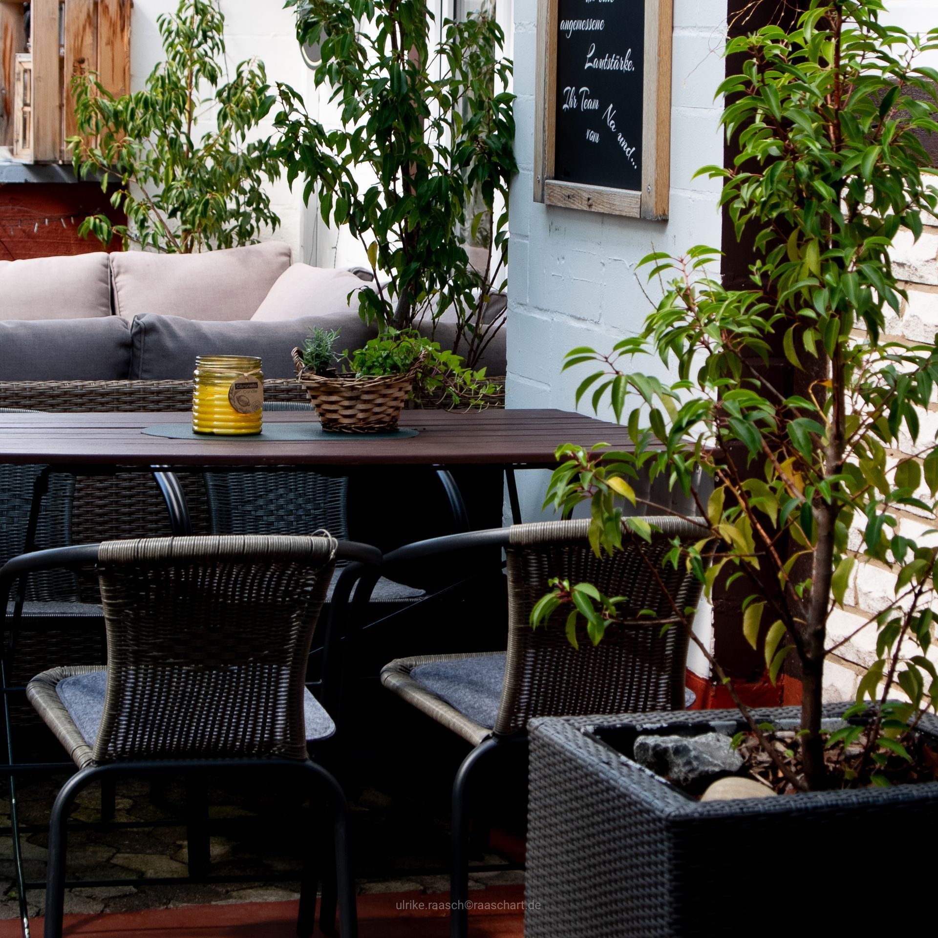 Terrasse im Freien mit Tisch und Stühlen, Pflanzen und einer Speisekarte auf einer Kreidetafel.