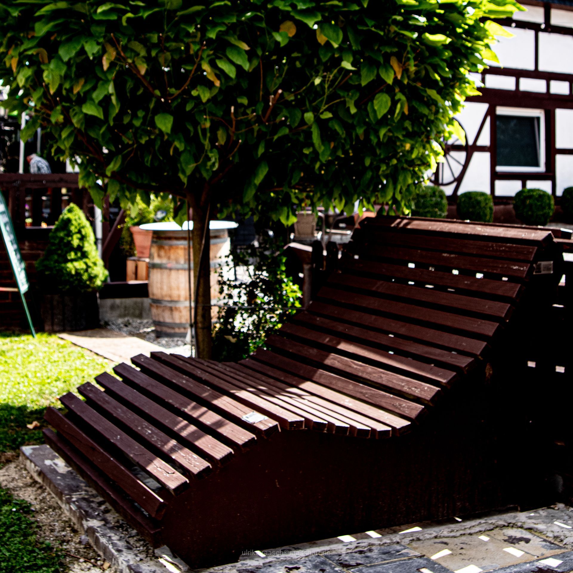 Eine hölzerne Chaiselongue unter einem belaubten Baum; im Hintergrund ein Fachwerkhaus.