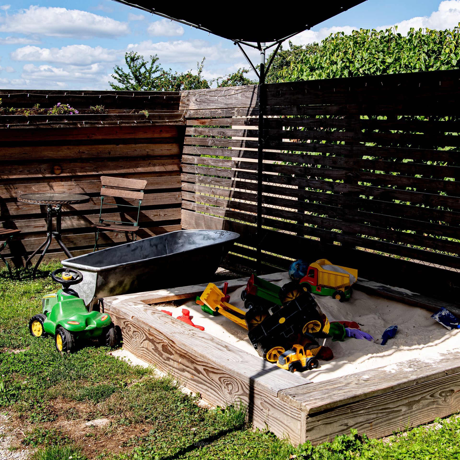 Ein Sandkasten mit Spielzeug, ein grüner Traktor und eine Metallwanne in einem Garten mit Holzzäunen und einem Sonnenschirm.