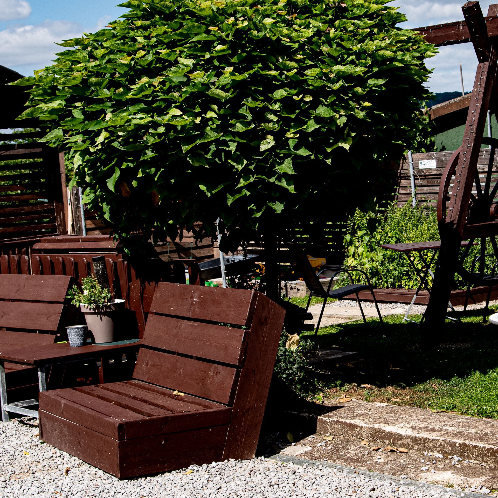 Braune Holzsitzgruppen im Freien in einem Garten mit großem, belaubtem Baum; Kiesboden.