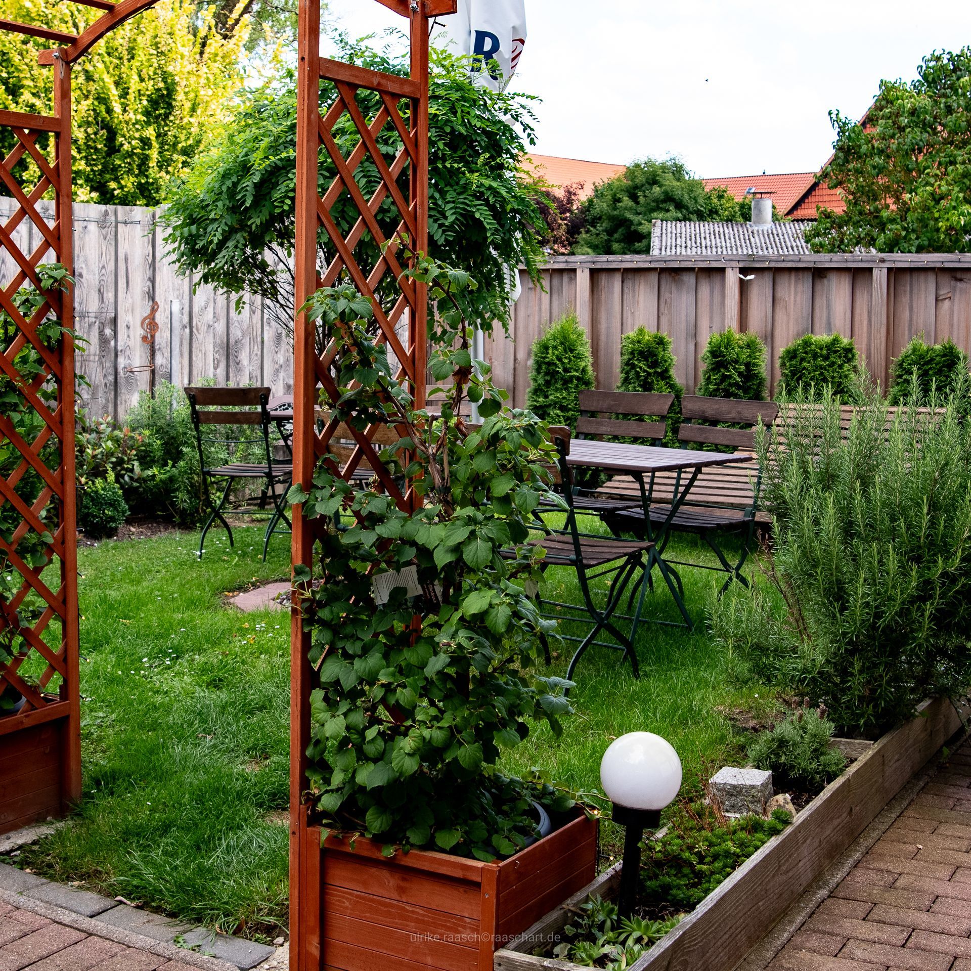 Hölzerner Garten mit Rankbögen, Tisch, Stühlen und verschiedenen Pflanzen.