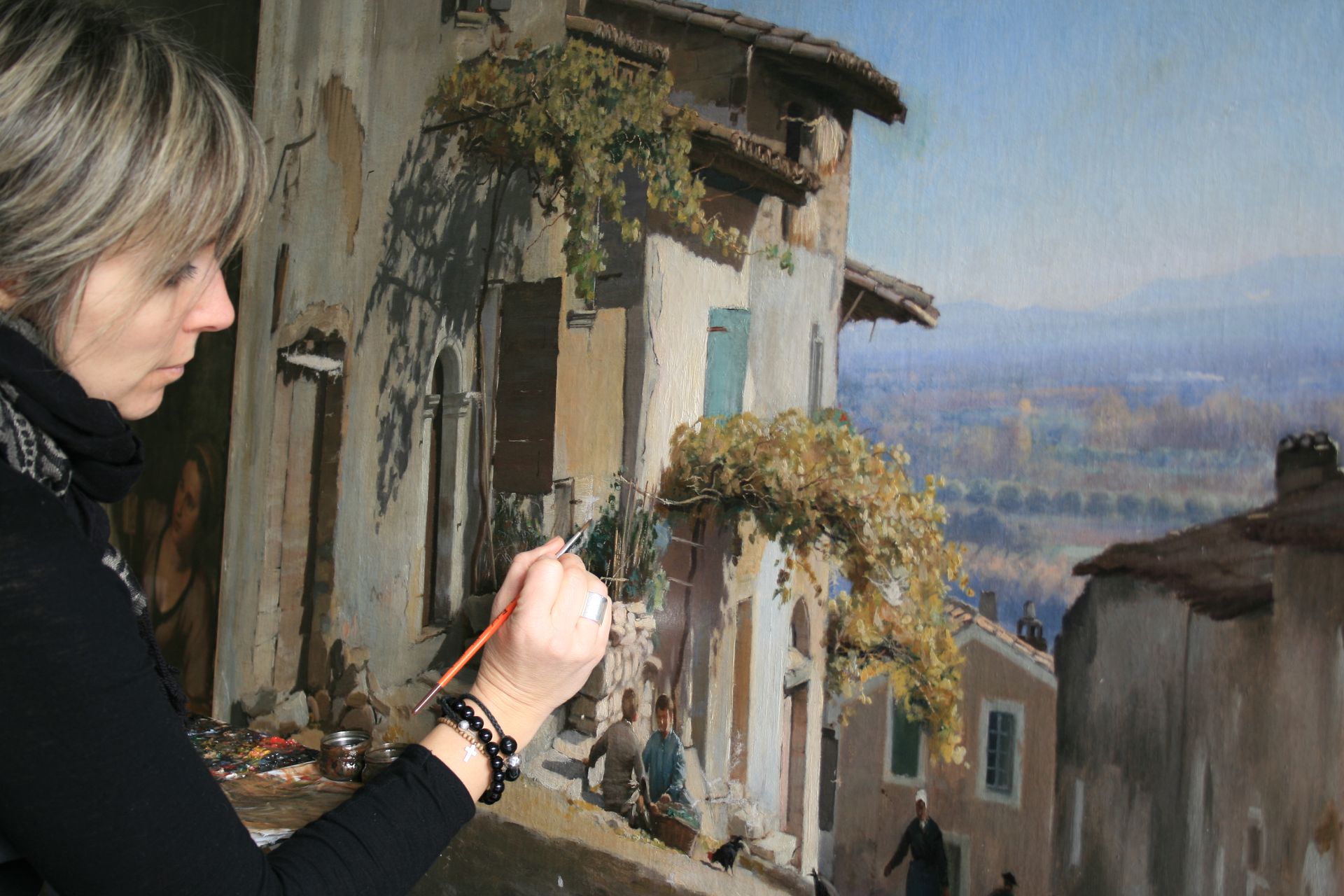 Restauratrice en train de rénover un tableau représentant des maisons.