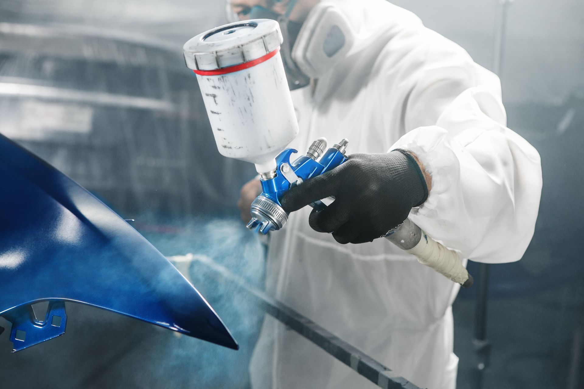 Un peintre carrossier applique de la peinture bleue sur une pièce de voiture dans un atelier, à l'aide d'un pistolet à peinture et d'une combinaison de protection.