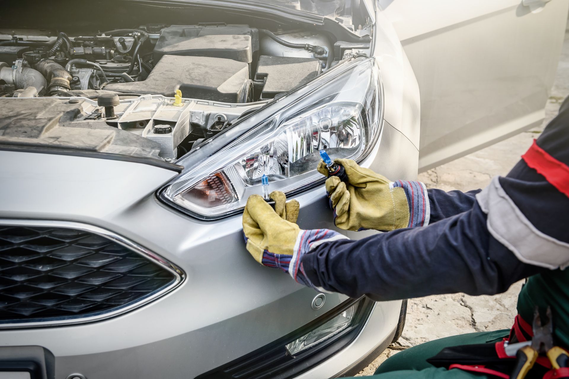 Un mécanicien portant des gants installe un phare sur une voiture argentée dans un atelier de réparation.