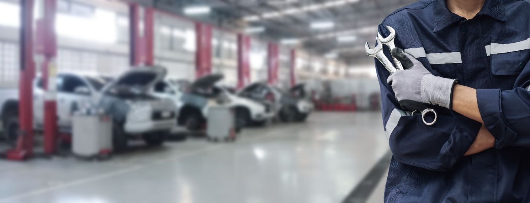 Un mécanicien, les bras croisés, se tient debout dans un garage automobile ; des voitures sont floues en arrière-plan.