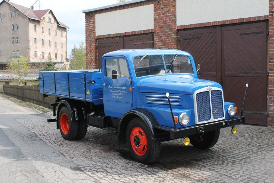 Blauer Oldtimer-Pritschenwagen parkt auf einer nassen Straße in der Nähe von Garagen und Wohnhäusern.