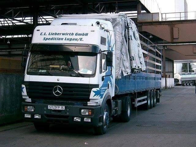Ein weißer Mercedes-Lkw mit blauen Seitenwänden parkt neben einem Industriegebäude unter einer Autobahnbrücke.