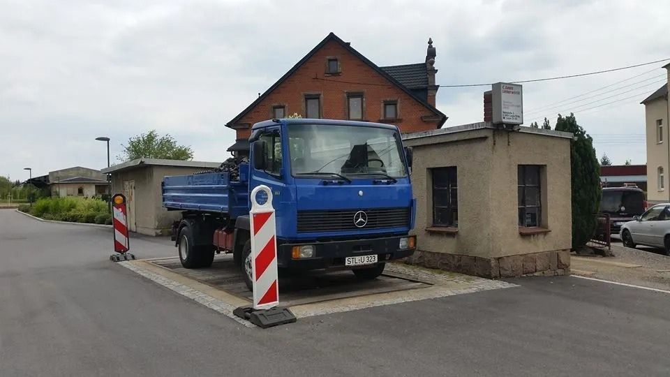 Ein blauer Muldenkipper steht neben einem kleinen Gebäude am Straßenrand, davor befinden sich rot-weiße Absperrungen.