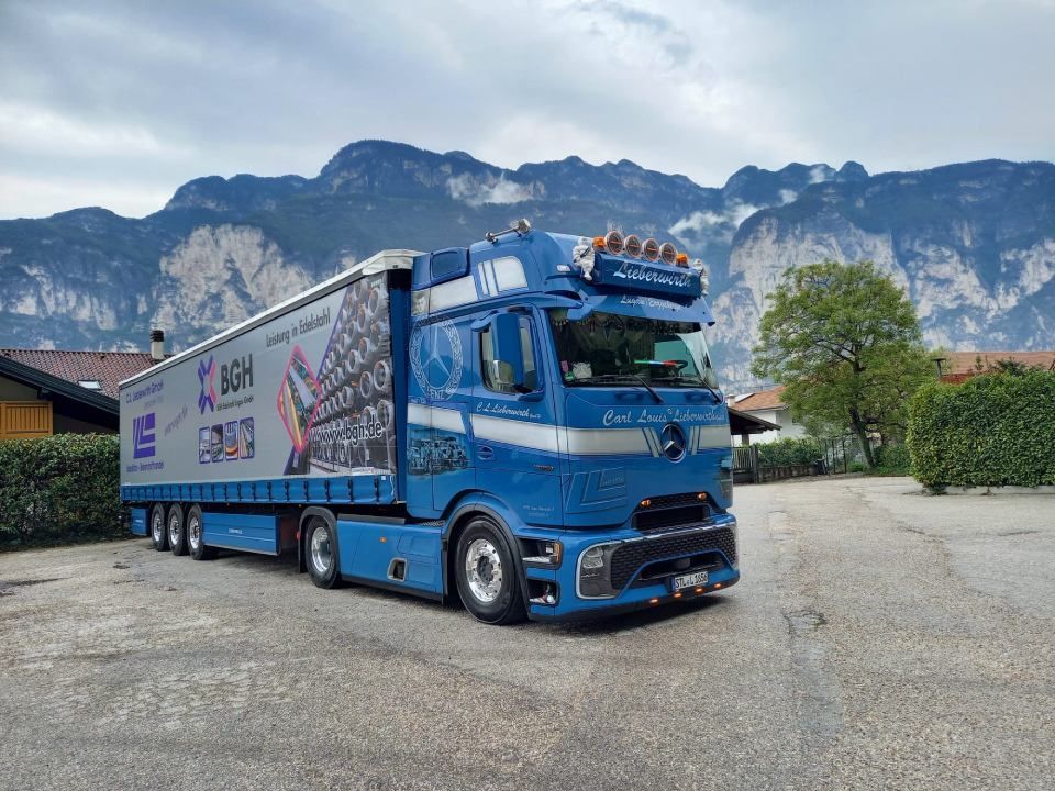 Ein blauer Sattelzug parkt auf Schotter vor einer Bergkulisse und bewölktem Himmel.