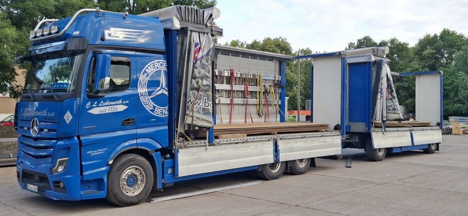 Blauer Lastwagen mit offenen Seitenwänden und Rampe parkt im Freien auf einem sonnigen Parkplatz.