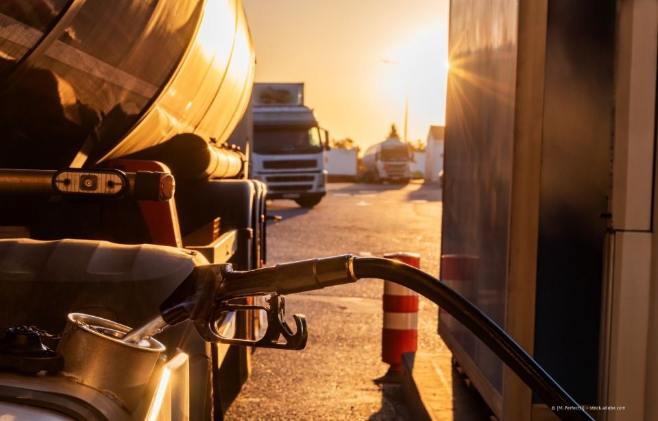 Eine Zapfpistole befüllt einen Tankwagen bei Sonnenuntergang; in der Nähe befinden sich orangefarbene Verkehrskegel und Industriefahrzeuge.