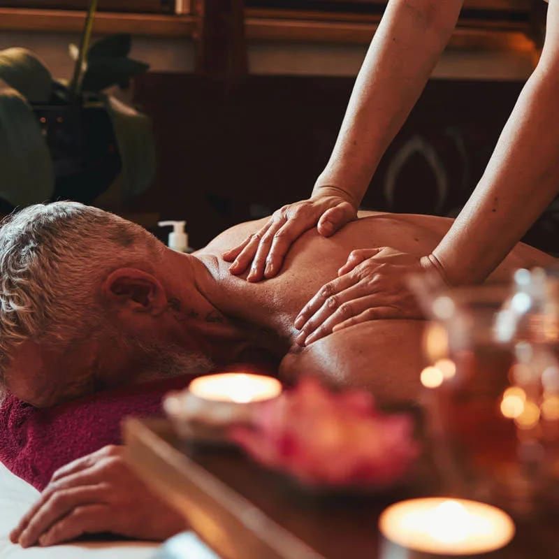 Persona recibiendo un masaje de espalda en un spa; manos masajeando, velas y piedras cerca.