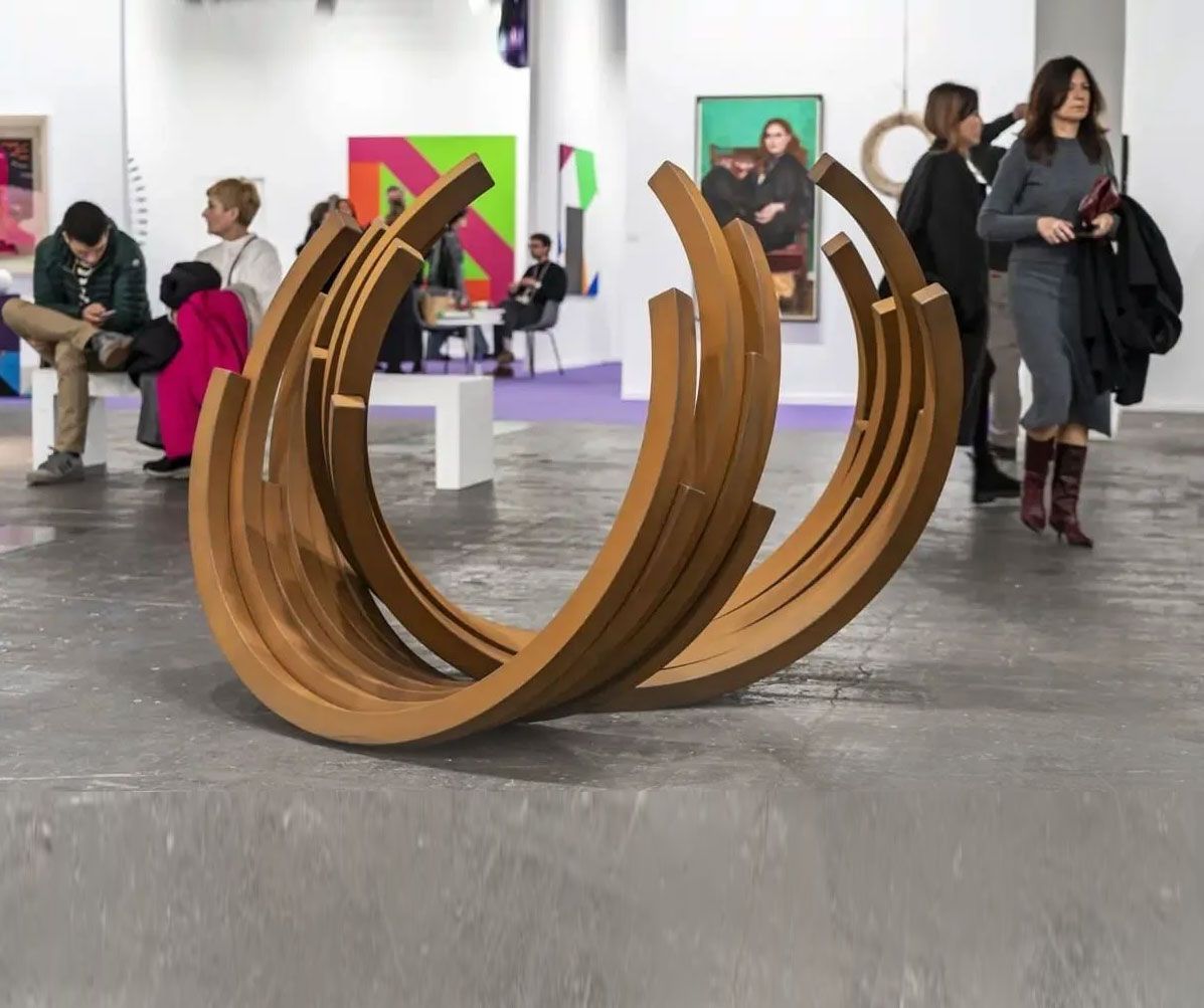 Escultura de madera curvada en una galería, con gente de pie y caminando a su alrededor en un espacio de exposición luminoso.