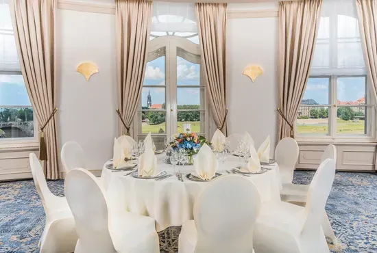 Lange, beige Vorhänge mit dekorativen Raffhaltern in einem hellen Saal mit hohen Bogenfenstern.