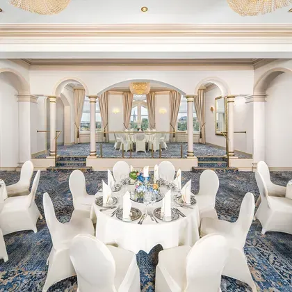 Weitläufiger Festsaal mit eleganten Säulen, runden Tischen und einer prachtvollen, hellen Deckengestaltung.